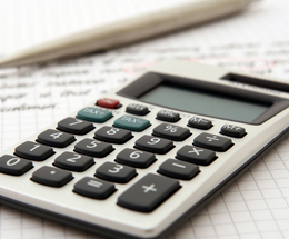 A calculator lays on a piece of paper with indistinct writing on next to a pen