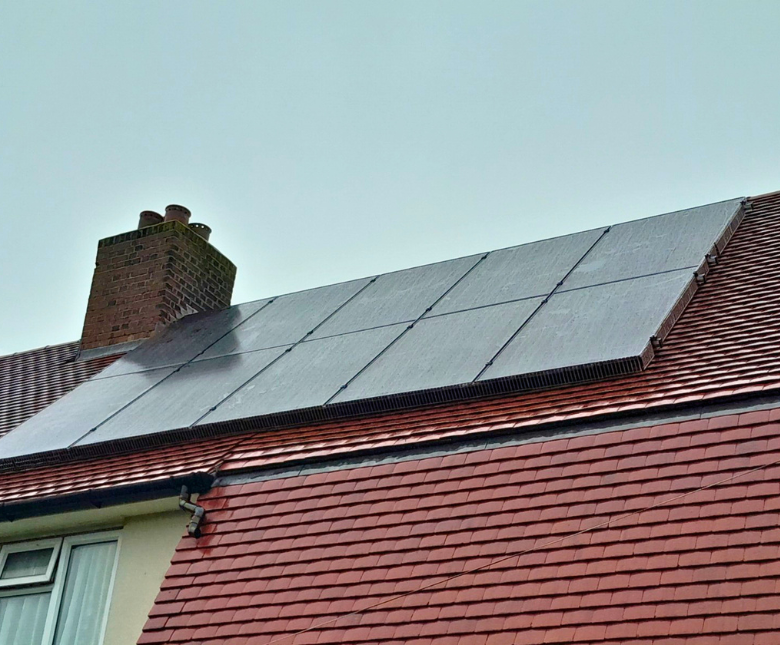 A residential house roof fitted with a row of large solar panels installed on dark mounting rails. The panels stretch across the upper slope of the roof beside a brick chimney stack. Below the panels, the roof is covered in red tiles, and part of a window
