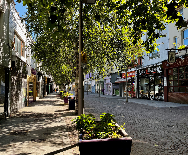 Tree-lined pedestrian shopping street with dappled sunlight filtering through leafy branches. Independent shops, including a barber shop and café, line the cobbled street, while large planters and seating areas create a welcoming town-centre atmosphere on