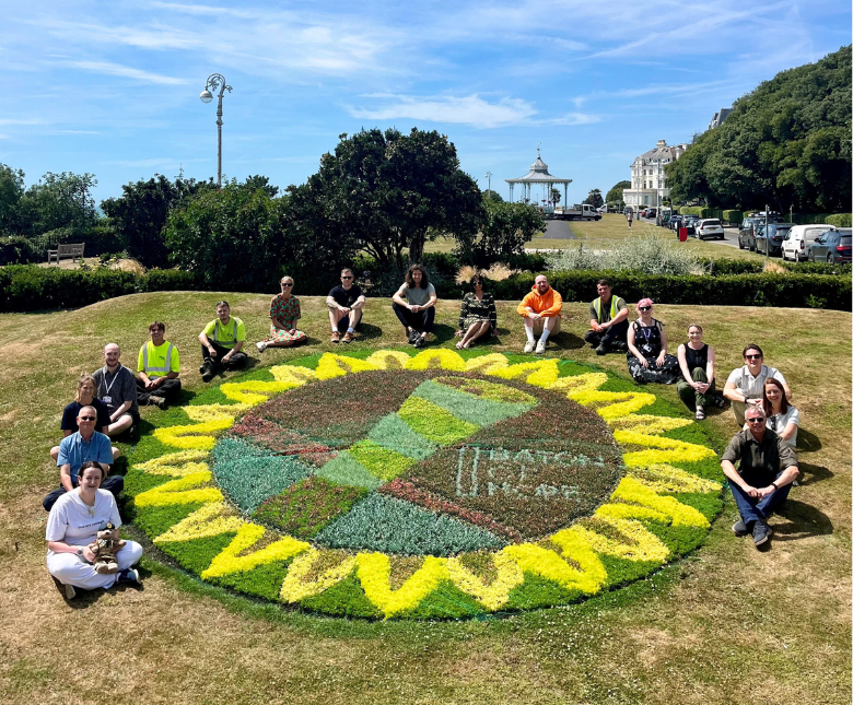 A large circular flower bed in Folkestone designed for the Baton of Hope campaign, featuring a green-and-brown awareness ribbon at the centre surrounded by bright yellow sunflower petals.