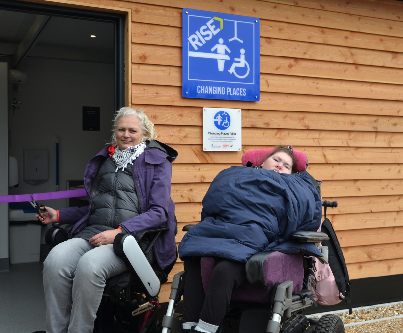 Two people in wheelchairs cutting a ribbon in front of the new Changing Places toilet in Folkestone.