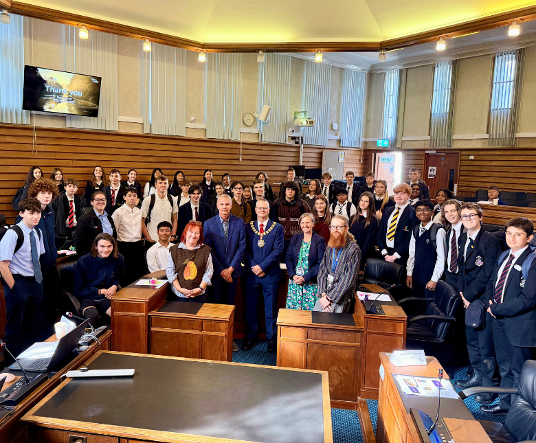A large group of secondary school and college students gathered in the council chamber at Folkestone Civic Centre for a Youth Forum event. The students, many wearing school uniforms, stand around the chamber alongside council representatives and community
