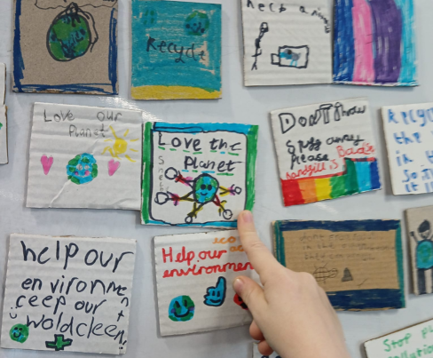 A young person's finger points to a noticeboard with messages urging loving the planet, recycling and helping the environment.