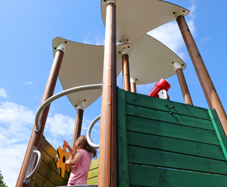 A low-angle view of a modern playground climbing structure with tall wooden posts and cream-coloured canopy roofs against a bright blue sky. Green wooden panels enclose part of the platform, while a red play telescope is mounted near the top.